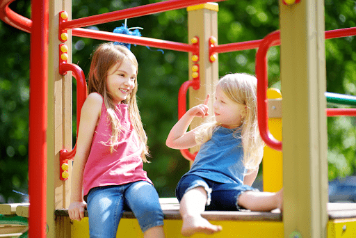 Two young girls sitting and playing together on a wooden playset.