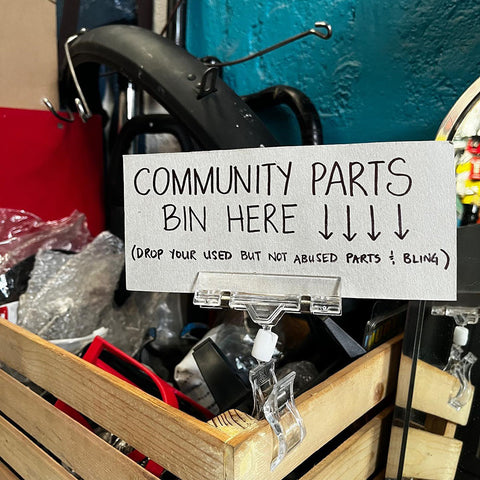 A basket full of donated bike parts for those in need with a sign saying "Community Parts Bin Here. Drop your used but not used parts and bling."
