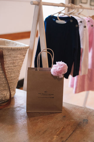 Sitting on a table you see a woven basket and a brown paper bag stamped with The Woolly Brand logo. It holds a merino wool blossom pink beanie with it's pom pom poking out the top. In the background some cable knit jumpers hang from a wooden frame.