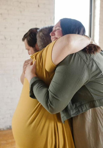 Photo is of Bethany and her husband Phillip hugging a family member. They are wearing greens, blacks and golden yellows in an industrial building.