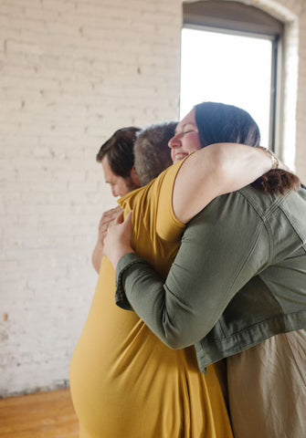 Photo is of Bethany and her husband Phillip hugging a family member. They are wearing greens, blacks and golden yellows in an industrial building.