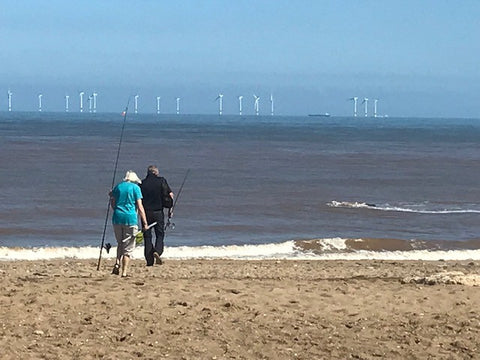 Wind turbines on the horizon of the sea. A couple walk toward the sea with fishing gear.