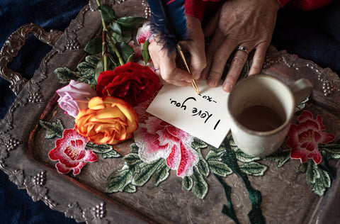 A grandma writting an "I love you note" on a metal tray with rose decorstions and a mug