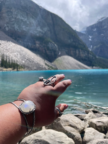 white rainbow moonstone cuff on wrist and sterling silver statement rings on fingers pointing at the canadian rockies at banff national park