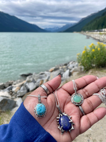 Turquoise stone sterling silver pendant, chrysoprase sterling silver pendant and lapis lazuli sterling silver pendant displayed on the palm of a hand in the lake and mountains backdrop