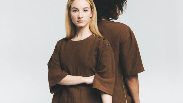 Two models stand back to back. The blonde woman faces forward while the brunette man has his back toward the camera. They are both wearing brown sustainable clothing against a white background.