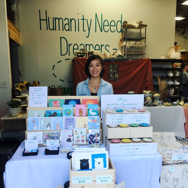 Table set up of cards and ceramics for sale and me (Chanamon) standing behind the table smiling.