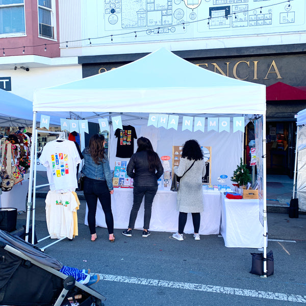 A photo of craft fair set up with a white tent and a few people looking at the merchandise.