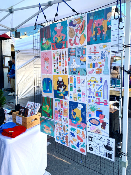 Photo of print display with art prints attached to a wire grid hanging from the tent.