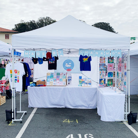 A photo of my booth set up with a white tent, signage, and colorful products.