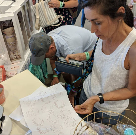 Woman stands in craft show line with hand drawn hairstyles deciding which doll to buy
