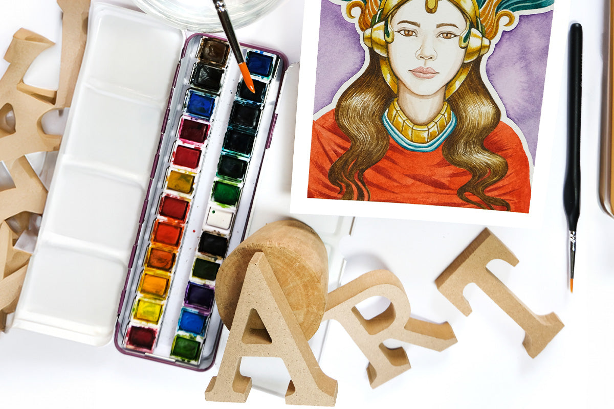 Flat lay photography of a watercolor painting, Artistry's artist-grade watercolor pan set, paintbrushes, and wooden alphabet letters spelling out "Art"