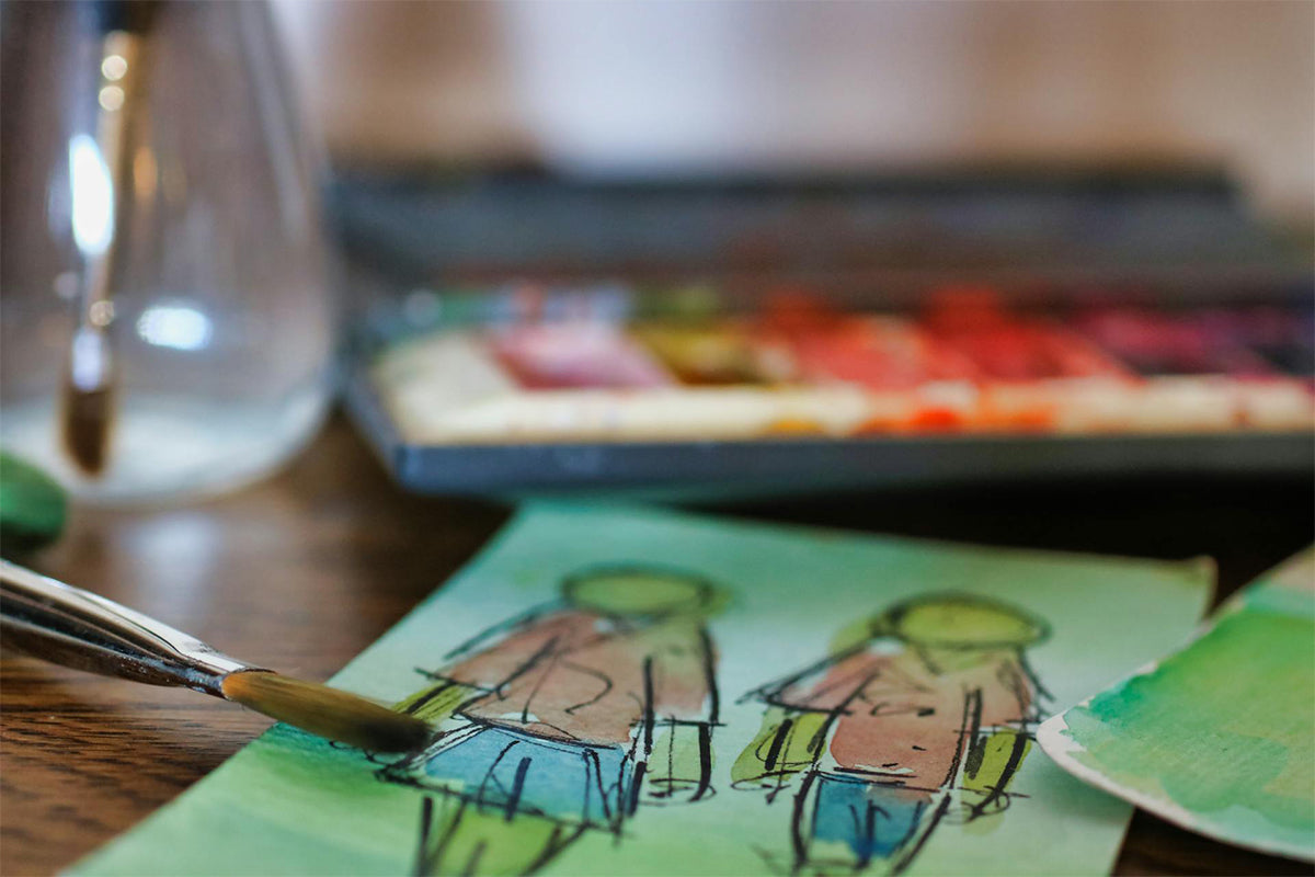A macro shot of a watercolor painting of a faceless girl and boy with a watercolor brush in the foreground