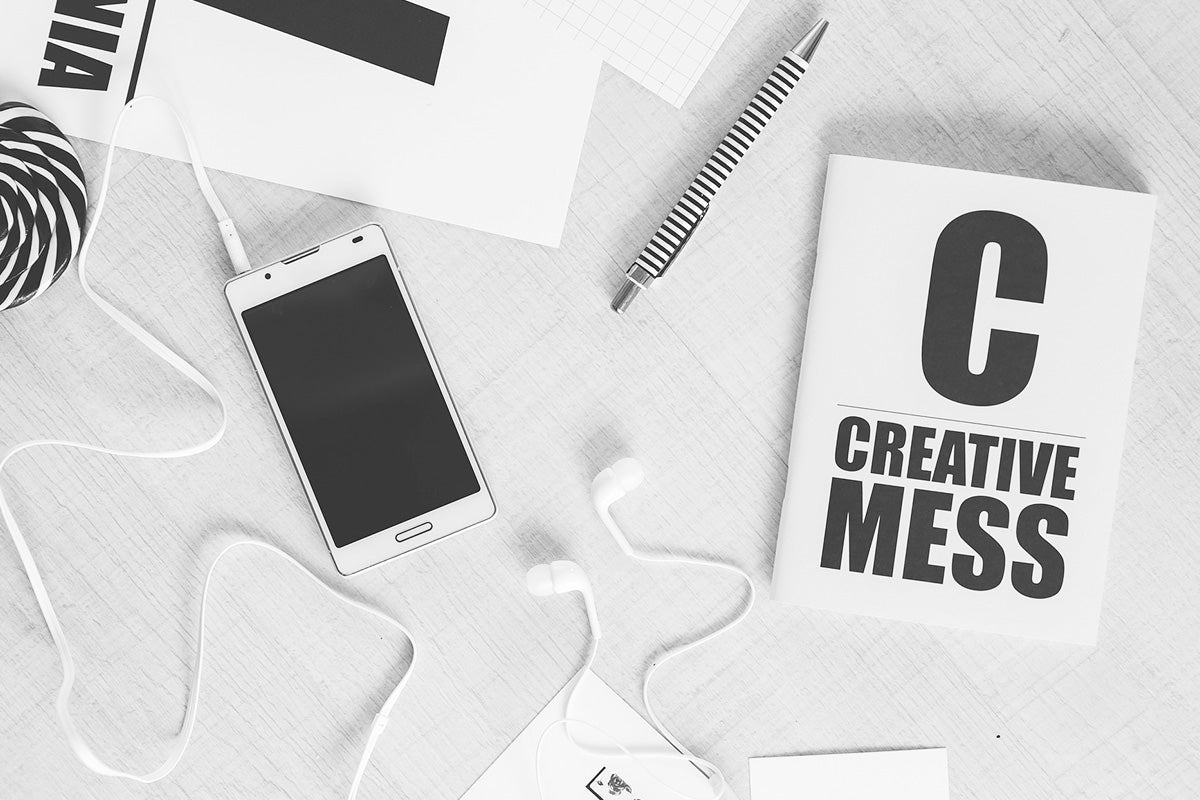 A black and white flat lay photo with a mobile phone, a pen, a pair of earphones, and a notebook with the print "Creative Mess" on it