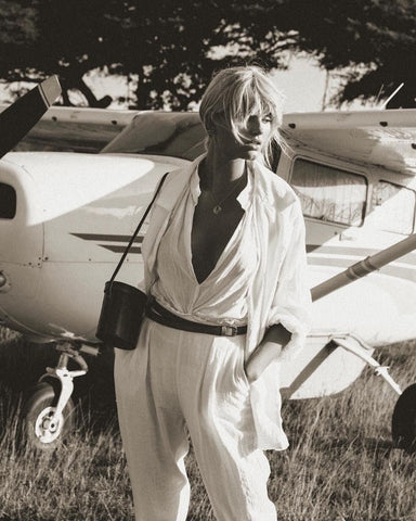 Black and white photo: Woman in all white linen pants and shirt standing in front of a small old plane.