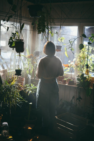 vrouw staat bij raam en kijkt naar buiten met hangplanten rond zich