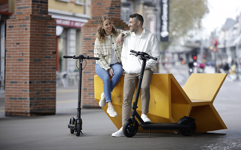 Ein junger Mann und eine junge Frau sitzen in einer Stadt lächelnd auf einer Mauer. Neben ihnen stehen zwei E-Scooter