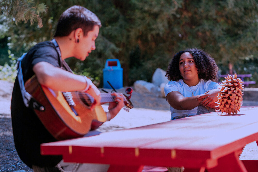 One Brave Trails Camper Plays Guitar While Another Watches At A Table