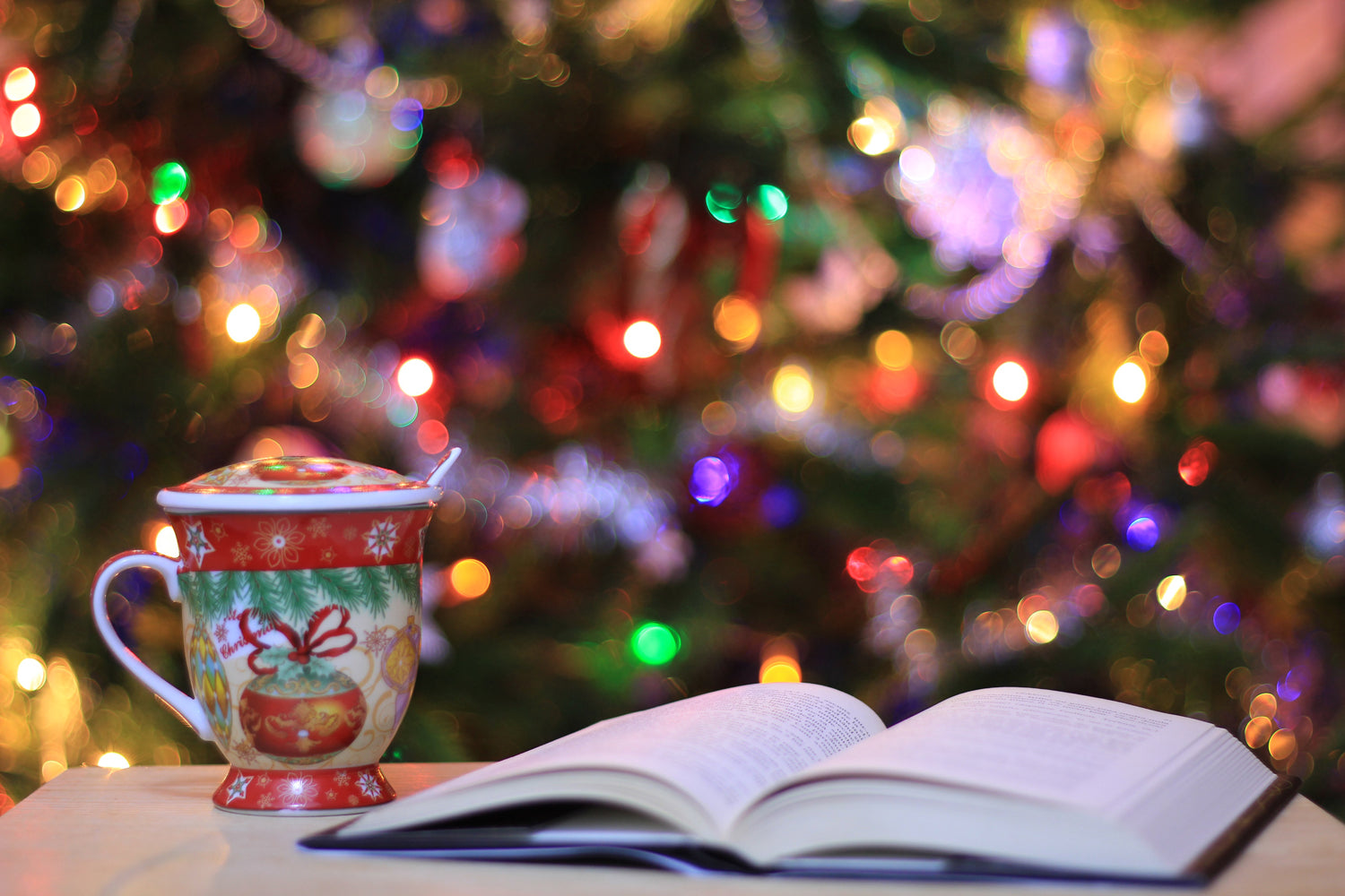 A Book And A Cup Of Tee On A Table With A Lit Up Christmas Tree And Lights In The Foreground