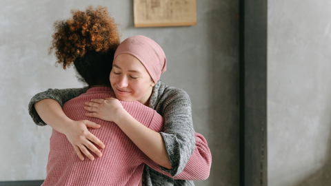 This is an image of two women hugging, one with her head wrapped in a pink cloth, and the other with a pink sweater, and super curly hair. This image is used in the Ippodaro Natural Salon blog post titled, “Why Hair Color Can Increase Your Risk of Cancer”.