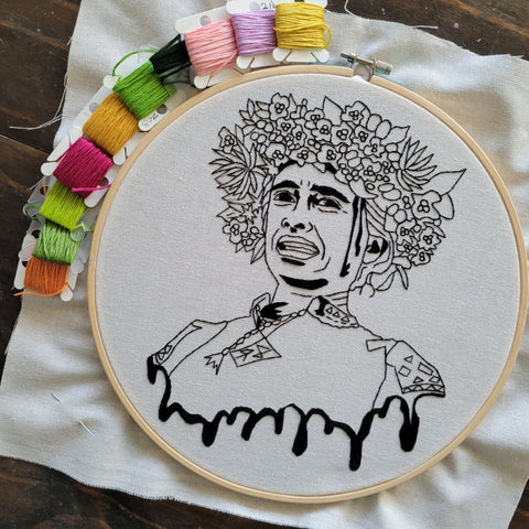 Midsommar (film) embroidery hoop with several bobbins of colourful thread surrounding it. The image on the hoop is outlined with a split backstitch and filled in some spots with the long and short stitch