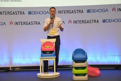 Wolfgang Höhn, der Gründer von Koru Kids und Koru Style, steht auf einer Bühne um das Unternehmen aus München, Bayern vorzustellen. Dabei stehen im Vordergrund ein paar Sitzherhöhungen von Koru Kids in verschiedenen Farben (rosa, blau, grün, grau). Dabei sind die Modelle Toddler Booster und Junior Booster