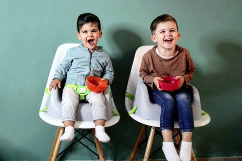 Zwei Kinder die auf dem Toddler Booster Grey mit einem 3-Punkt-Haltegurt sitzen, welcher eine Sitzerhöhung von Koru Kids ist