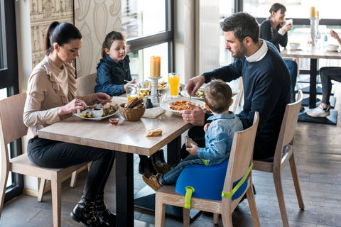 Eine Familie wie sie zusammen an einem Tisch isst und dabei ein Kind, das auf einem blauen Toddler Booster von Koru Kids sitz. Toddler Booster ist eine Sitzerhöhung