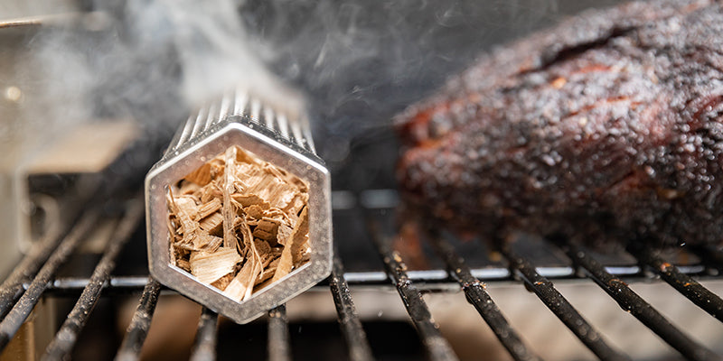 Smoker tube filled with smoking wood chips next to smoked BBQ meat on a grill