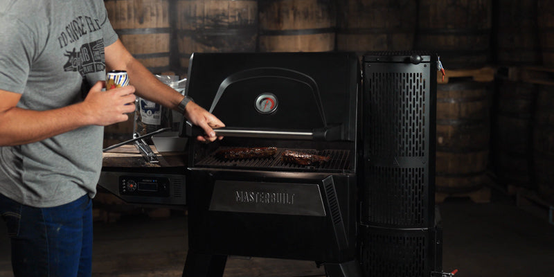 A man holding a barrel-aged beer, lifting the lid of a smoker with meat inside and used bourbon barrels in the background