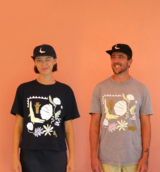 Sarana and Tom stand against an orange background. They are wearing matching Aslan Ruby Studio tees and matching Aslan Ruby Studio hats that are black with a pink crocodile embroidered on them. Tom is smiling like he's accomplished something pretty great. Sarana is squinting with one eye shut because sometimes she feels uncomfortable smiling at the camera in photoshoots and for some reason she feels this is a better way to present herself.