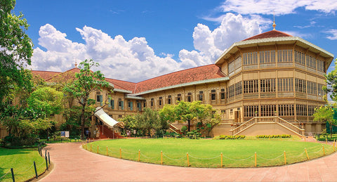 Vimanmek Palace in Thailand, a majestic all-teak construction with a multi-tiered roof and intricate wooden facade, set against a lush garden and clear blue sky.