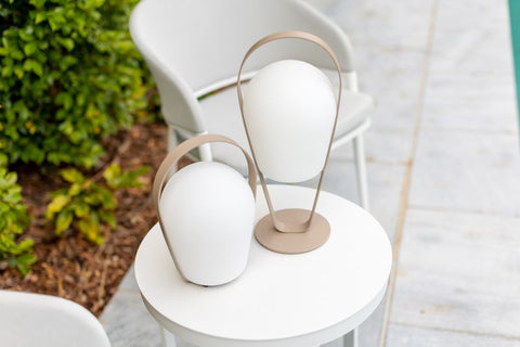 Remarkable Outdoor Living: Bobby Outdoor Aluminium Lamp Low.This image features two modern portable LED lamps placed on a round white table. The lamps have a minimalist design, with matte white diffusers and a sleek bronze-coloured handle that curves around the diffuser. The handle serves both as a design element and a functional part, making it easy to carry the lamps. The white and bronze colours suggest a contemporary and elegant aesthetic, fitting well for modern outdoor or indoor living spaces. The greenery in the background provides a nice contrast and indicates that this setting is outdoors, perhaps on a patio or deck.