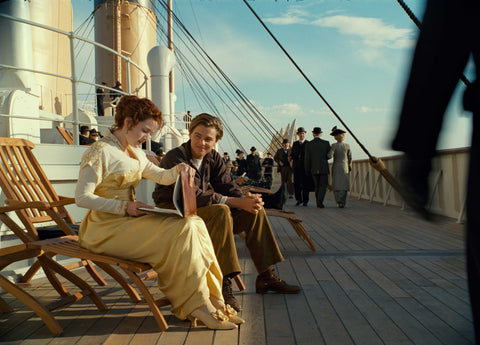 A still from a movie set aboard the historical RMS Titanic, showing a young couple seated on wooden deck chairs. The deck is constructed from teak wood, which shines under the daylight, emphasising the luxury of the ship.