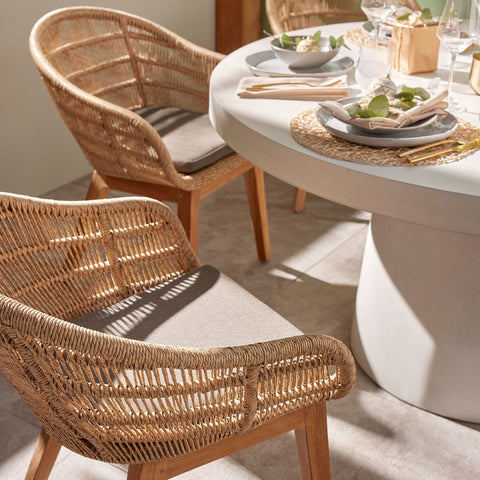 Zen Round Table Monsoon Chair Outdoor Dining Setting featuring wicker chairs with plush cushions and a round, white dining table set with plates and cutlery. The setting is in a sunlit area casting soft shadows.