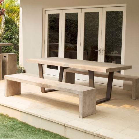 A Zen Outdoor Dining Setting showcasing a rectangular concrete table and two matching benches, positioned on a pale tiled patio and bathed in soft sunlight. It is bordered by lush green grass on one side and a view of a glass door that leads into a home, hinting at an indoor-outdoor living space.