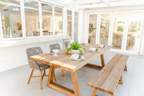 Valley Outdoor Recycled Teak Dining Table, Bench, and Armenia White Wicker Dining Chairs showcased in a bright outdoor dining area. Rustic wooden furniture, white dishes, and a central plant create a clean ambiance, complemented by natural light.