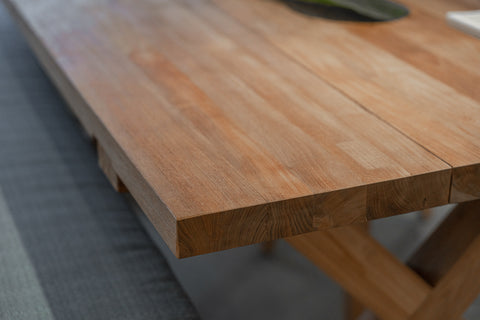 Bunbury Outdoor Recycled Teak Dining Table close-up, revealing a smooth, warm-toned wooden surface with natural grain. The sturdy construction and quality craftsmanship are emphasised from a side angle, set against a soft, textured grey background.