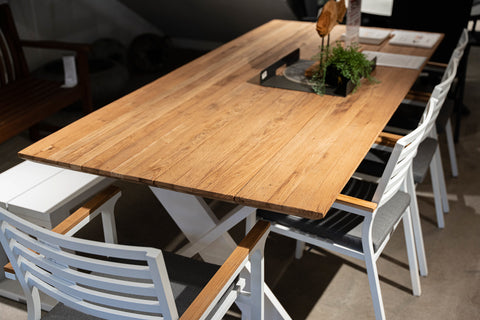 Carmel Outdoor Recycled Teak Dining Table in White showcased in an indoor setting, with a natural finish, paired with modern white chairs with grey seat cushions and teak arms. A central cut-out holds decorative green plants, adding a unique touch.