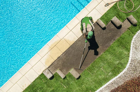 An aerial view of a person in a green shirt sweeping a pathway beside a bright blue swimming pool, with neatly landscaped grass and garden details around.