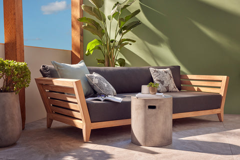 The image depicts a tranquil patio setting, showcasing an Ottawa Teak Daybed with comfortable cushions and a Zen Concrete Round Stool serving as a side table, all complementing a Mediterranean aesthetic.