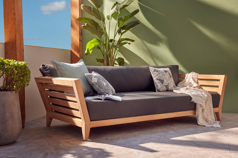 Ottawa II Outdoor Teak Daybed features a modern design with charcoal cushions, accented by light grey and patterned throw pillows, and a cozy beige blanket. Set against a green wall and large leafy plant, creating a tranquil outdoor ambiance.