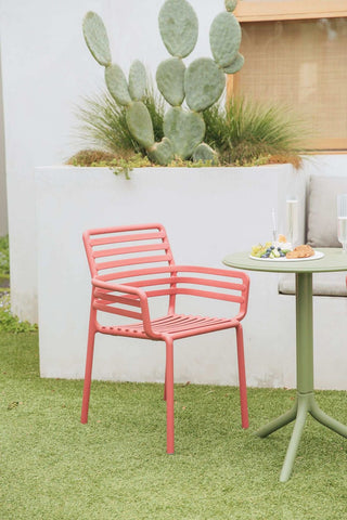 Remarkable Outdoor Living: Nadi Doga Outdoor Resin Dining Armchair. The image presents a neatly composed outdoor setting featuring a coral pink chair with a simple, modern design. The chair is accompanied by a round table with an olive green stand, upon which rests a small plate of cookies, a bunch of grapes, and two elegant champagne glasses, suggesting a prepared snack or light refreshment. In the background, a large cactus within a white raised planter adds a touch of natural greenery to the scene, which contrasts nicely with the chair and table colours.