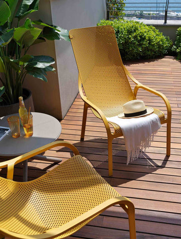 Remarkable Outdoor Living. Nardi Net Outdoor Resin Balcony Lounge Chair. This image shows a vibrant outdoor setting with a yellow perforated metal lounge chair and matching table set against a backdrop of lush greenery and a clear sky. A white hat with a black band rests on the chair, adding a leisurely touch to the scene. The ensemble is situated on a wooden deck, enhancing the warm and inviting ambiance perfect for relaxation or socialising outdoors.