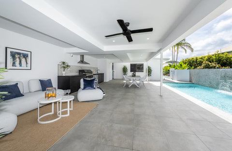 This image features a sleek, outdoor poolside lounge area with a modern Iowa Lounge sofa with blue cushions and a Neverland side table in white. A grey-tiled patio holds the furniture, while a tranquil pool with a waterfall edge and tropical foliage completes the serene setting. A black ceiling fan above adds comfort to this luxurious relaxation spot.