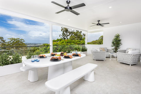 This image displays a pristine white Bristol dining set, including a modern table and matching bench, positioned on an open balcony. The dining surface is adorned with place settings for a meal. In the background, cozy Lawson lounge chairs invite relaxation, overlooking a breathtaking view of a lush landscape and distant horizon under a clear sky, with a ceiling fan above adding a touch of indoor comfort outdoors.