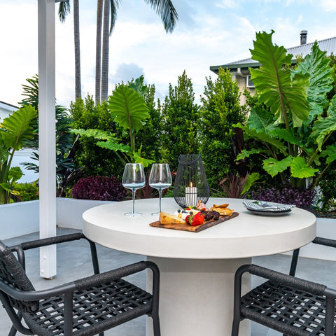 This image captures a Zen outdoor concrete table on a balcony, paired with charcoal Helena rope chairs. On the table, there's a wooden platter with snacks and two wine glasses, accentuated by a candle lantern. Lush greenery surrounds the intimate setting.