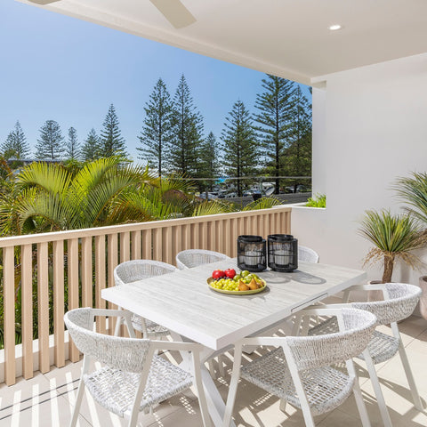 This image depicts a Clifton Outdoor Extension Table accompanied by Auto wicker chairs, both in White, on a sunny balcony. The table is set with a bowl of fresh fruit and two black lanterns, against a backdrop of tall pine trees and lush palms, capturing a relaxed dining ambiance.