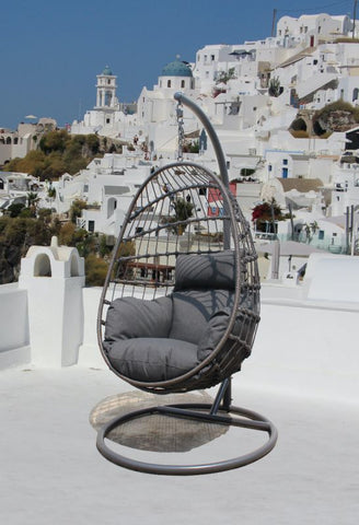 The Dayton Outdoor Rope Hanging Egg Chair with gray cushions, situated on a white terrace overlooking the classic white architecture and blue domes of Santorini, Greece.