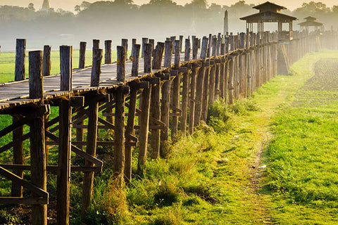 The U-Bein Bridge, a long wooden footbridge made of teak, stretches across the landscape at sunrise, with a dirt path running alongside it.